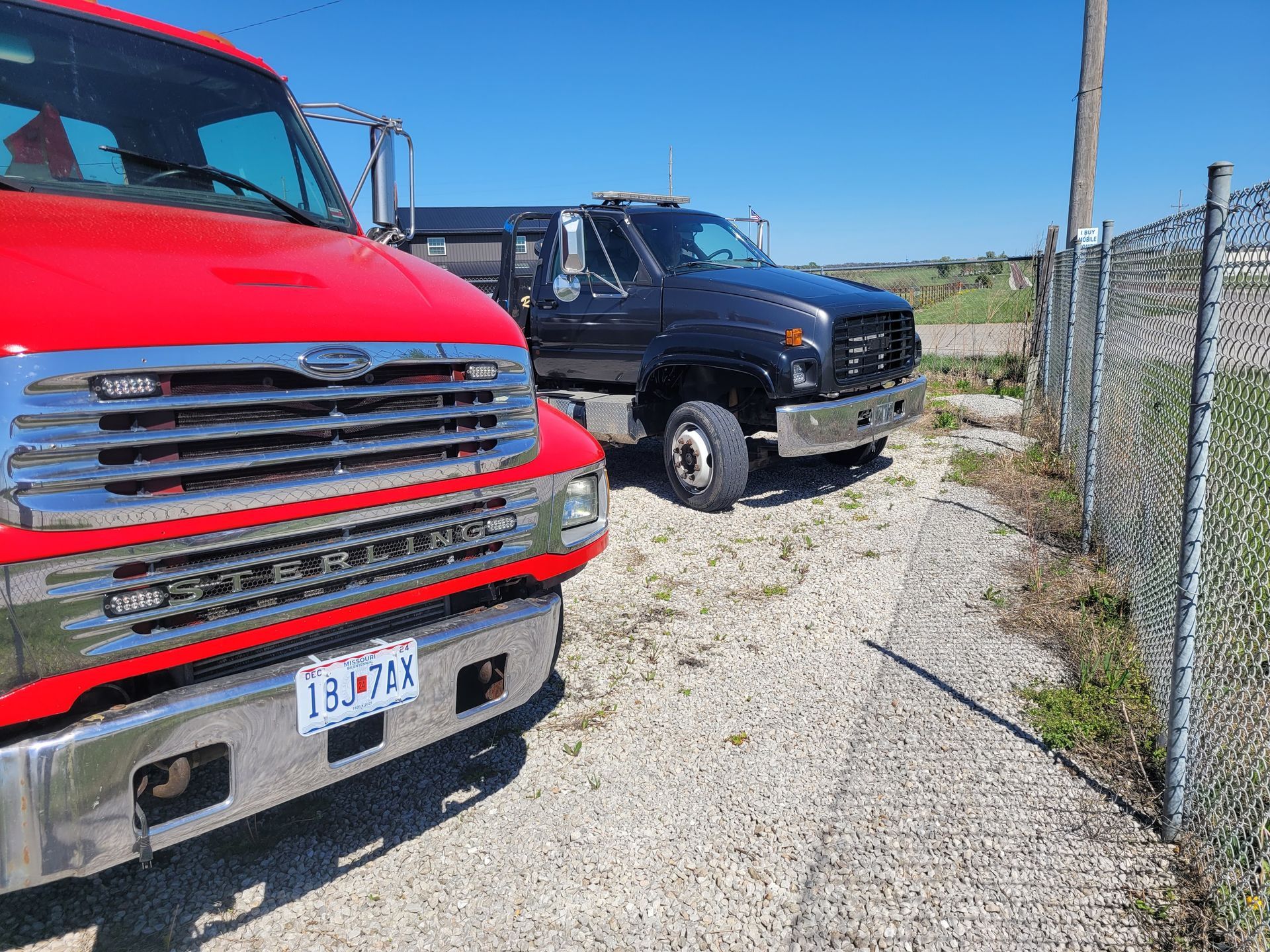 Red and black work trucks parked side-by-side on a gravel lot next to a chain-link fence on a sunny day.