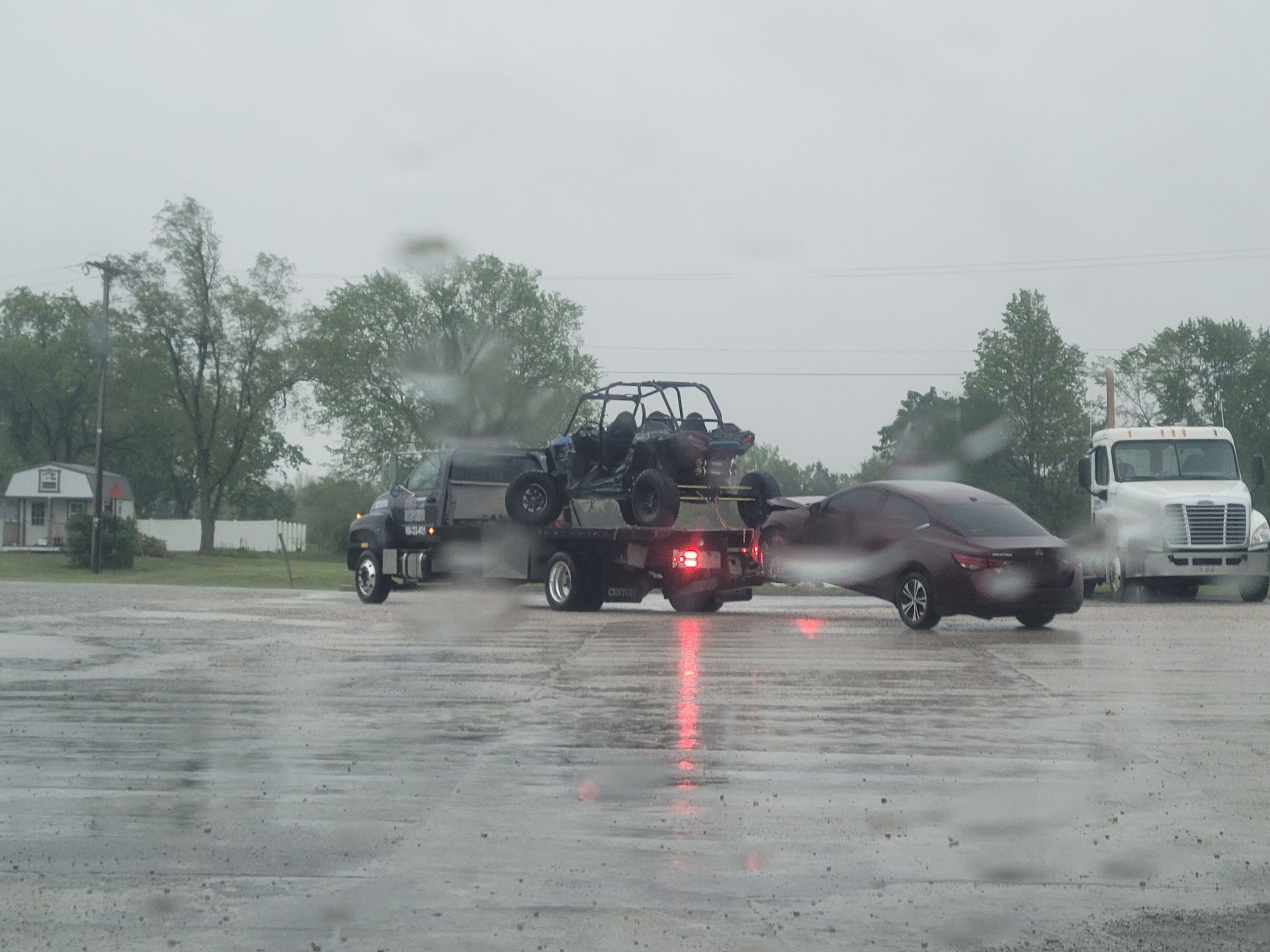 Truck hauling an ATV and a car on a rainy day in a parking lot.