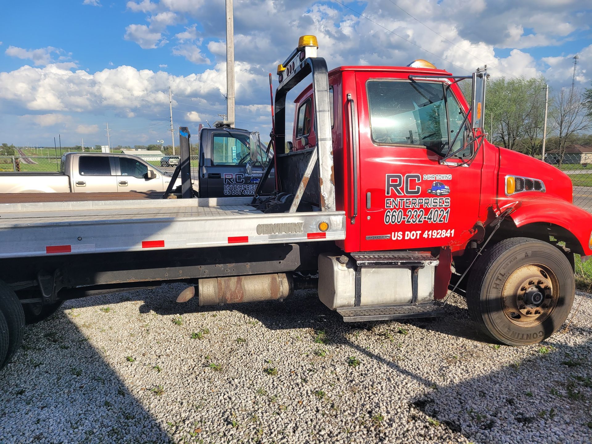 Red tow truck with a flatbed parked on gravel. In the background, a white truck and blue sky.