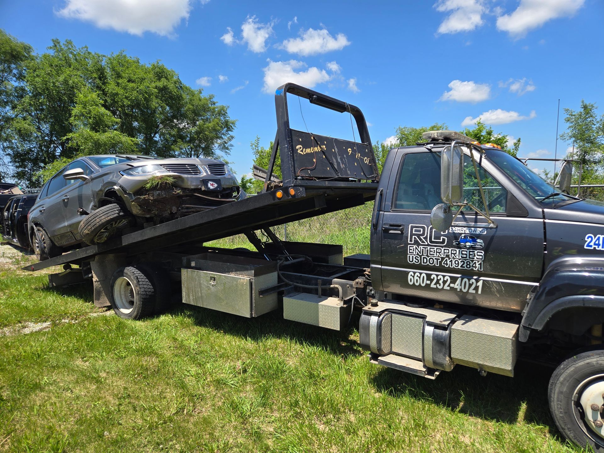 Gray car on a flatbed tow truck in a grassy area on a sunny day.