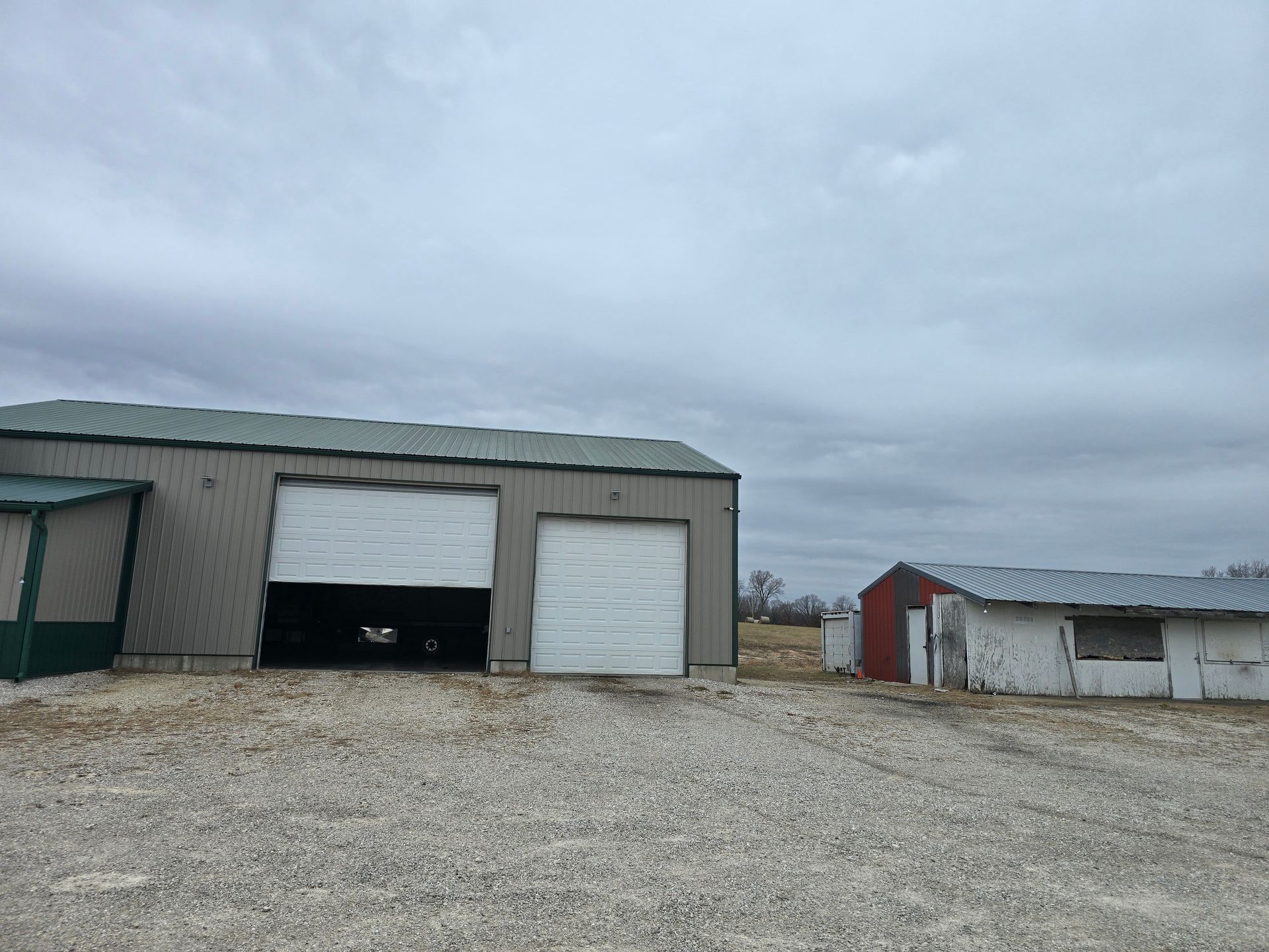 Gray building with open garage door, small red shed, gravel lot under cloudy sky.