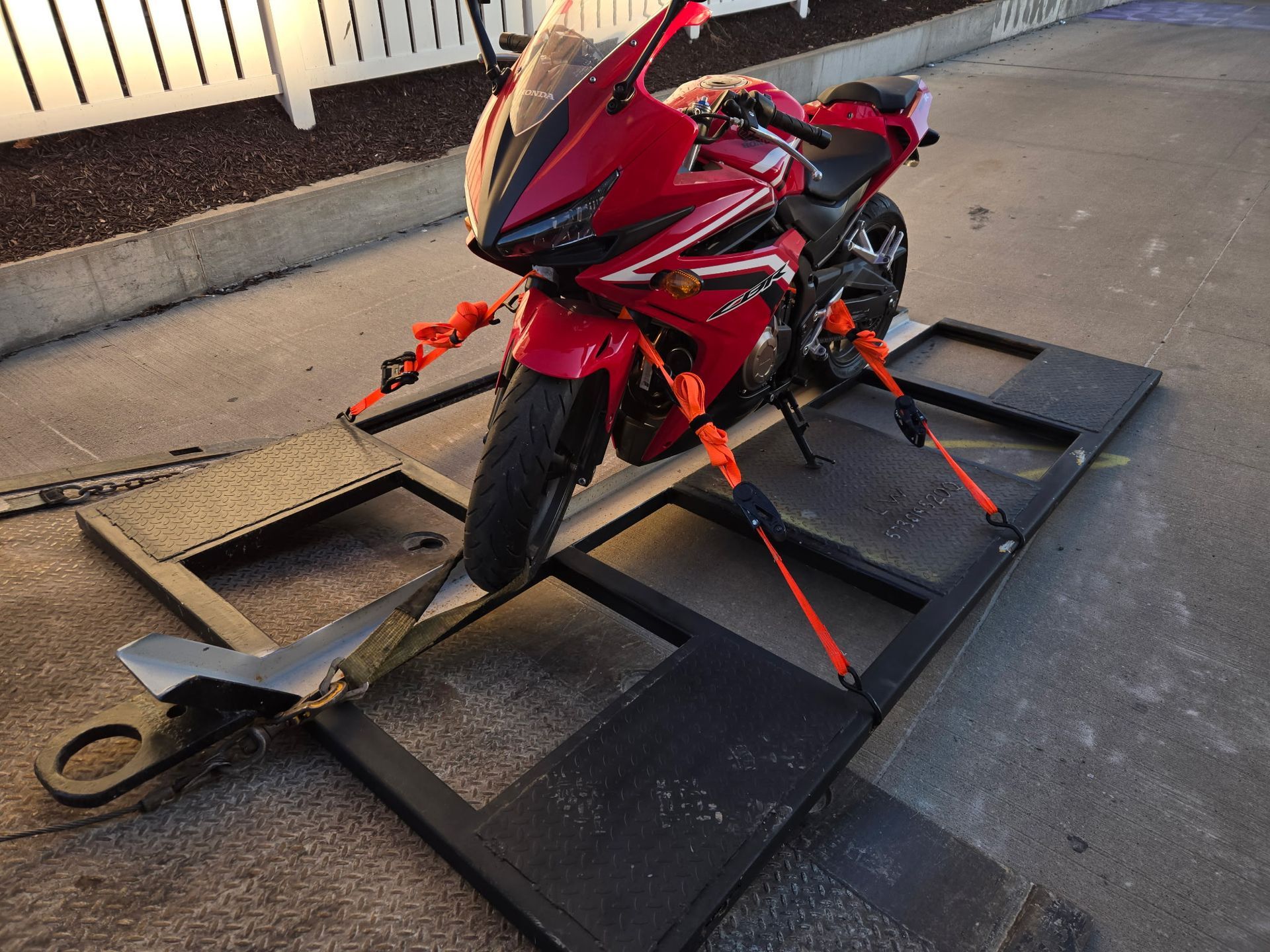 Red motorcycle secured to a black trailer with orange straps. Outdoors, parked near a building.