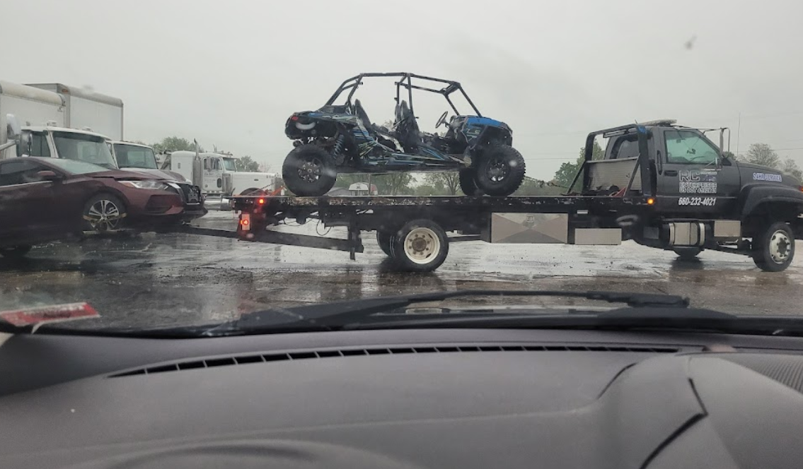 Tow truck carrying a damaged off-road vehicle on a rainy day; car in tow behind.