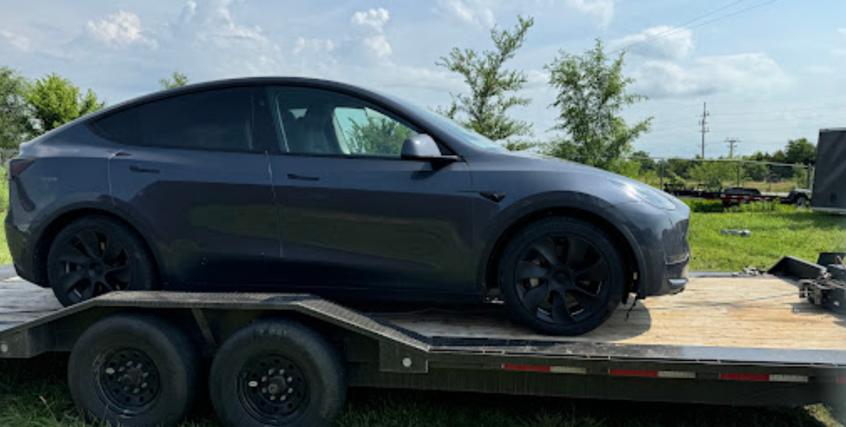 Dark gray Tesla Model Y on a trailer in a grassy outdoor setting.
