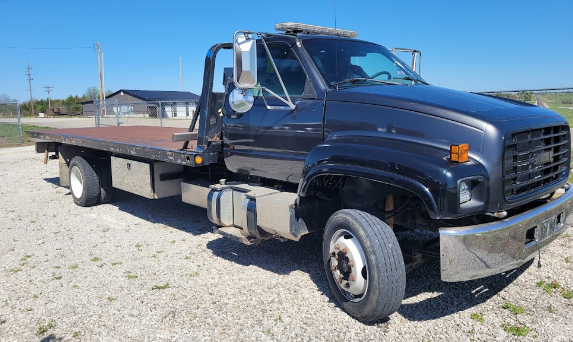 Black flatbed tow truck on a gravel lot under a clear blue sky.