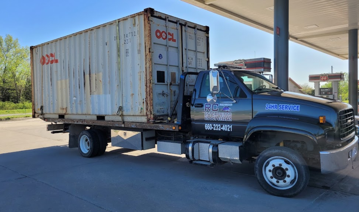 A large truck with a shipping container on its back parked under a canopy at a gas station.