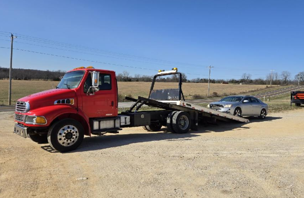 Red tow truck with a silver car on its flatbed, parked outdoors on gravel.