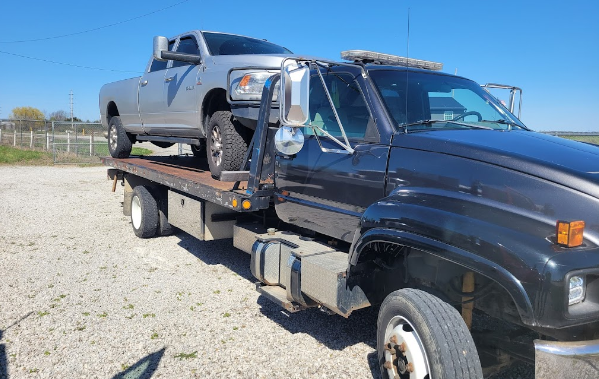 A silver pickup truck loaded on a black tow truck, parked outdoors on a sunny day.