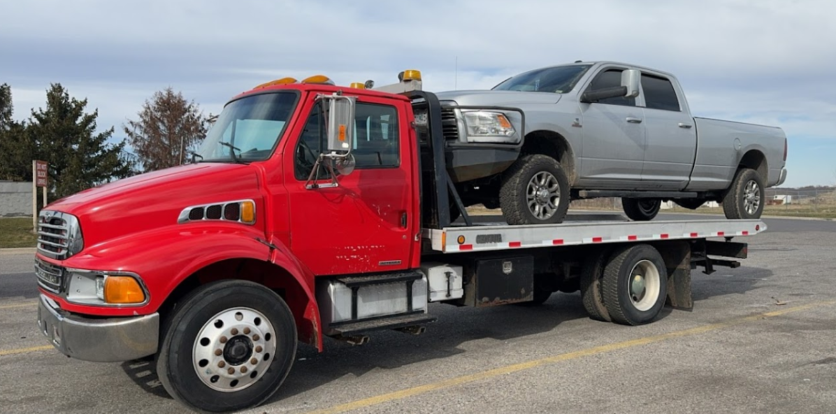 Red tow truck transporting a gray pickup truck on a flatbed. Outdoors, cloudy sky.