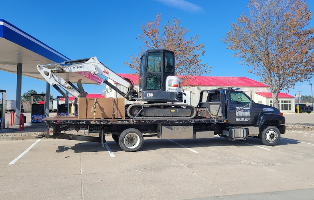 A Bobcat excavator on a flatbed truck at a gas station, blue sky.