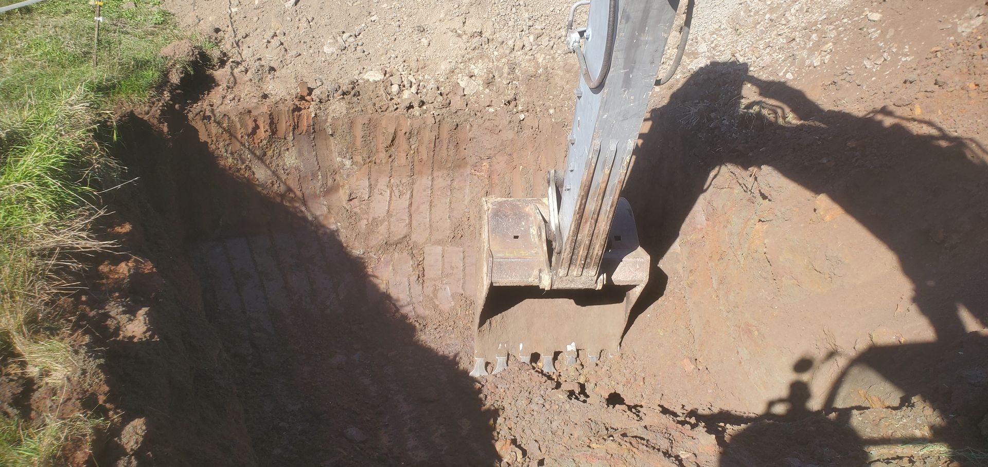An excavator digging a trench in the dirt, casting a long shadow. The edge of a grassy area is visible on the left.