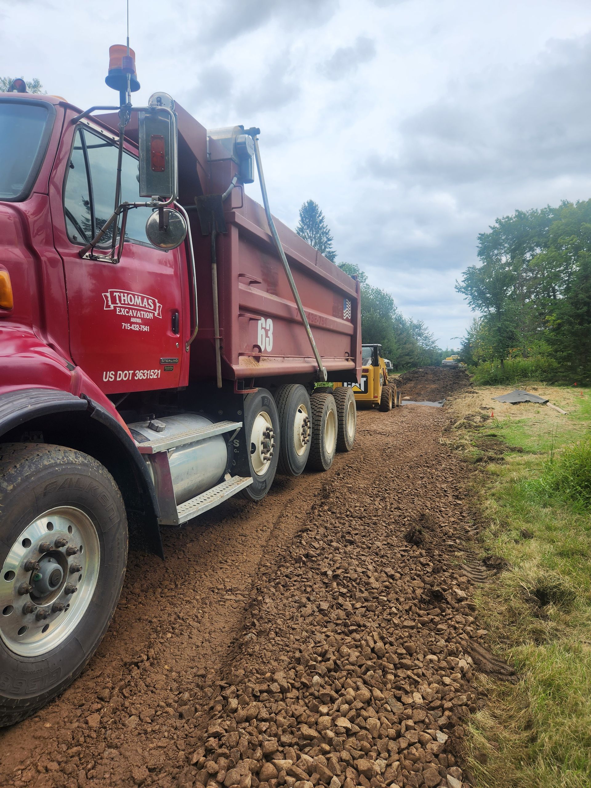 Red dump truck on a gravel road, unloading material. A yellow front-end loader sits in the background, forest on the right.