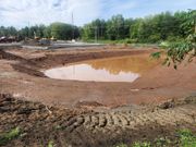 A muddy pond surrounded by red dirt is reflecting the sky. Construction equipment and trees are in the background.