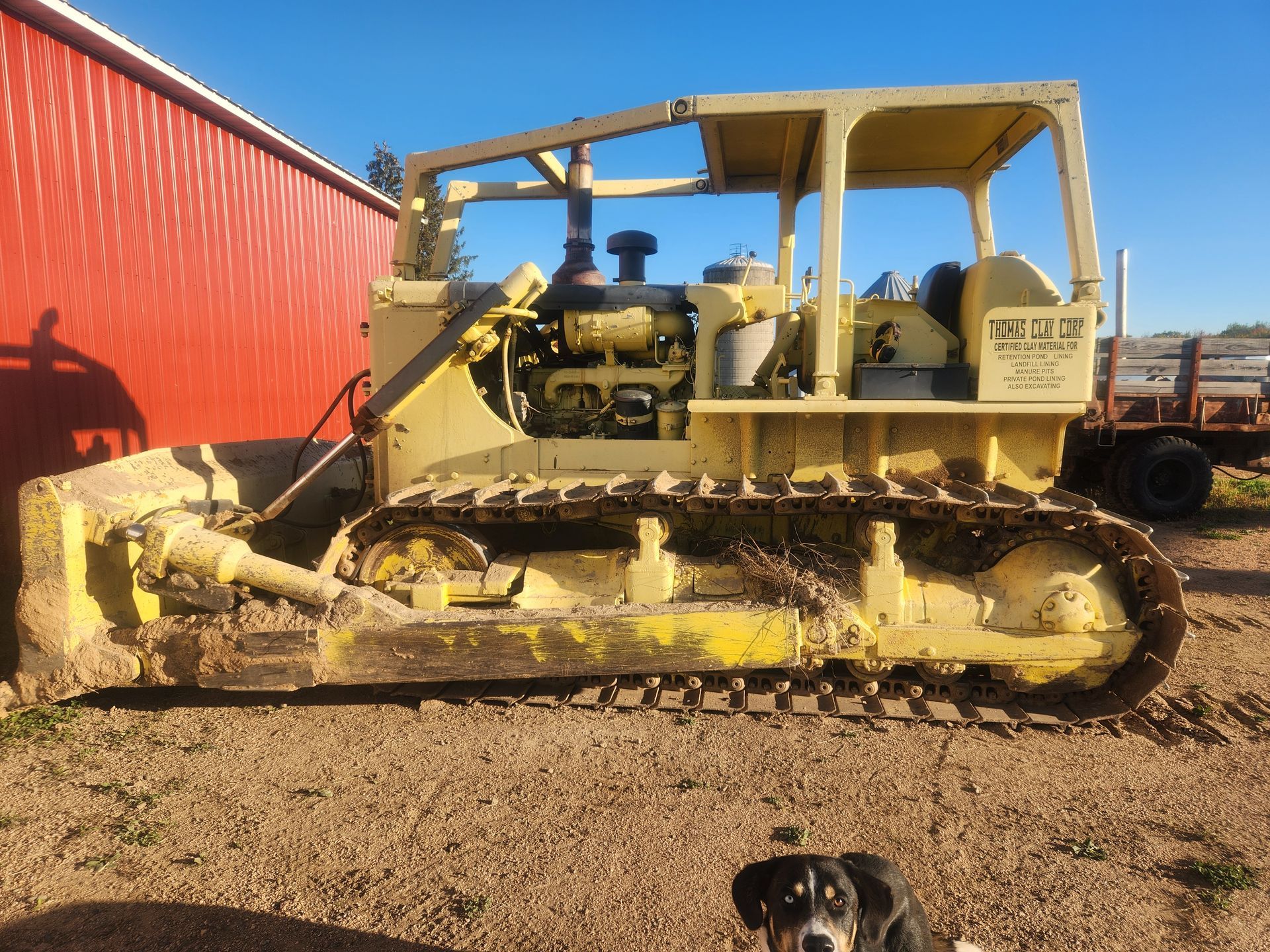 Yellow bulldozer parked in front of a red building; dog in foreground.