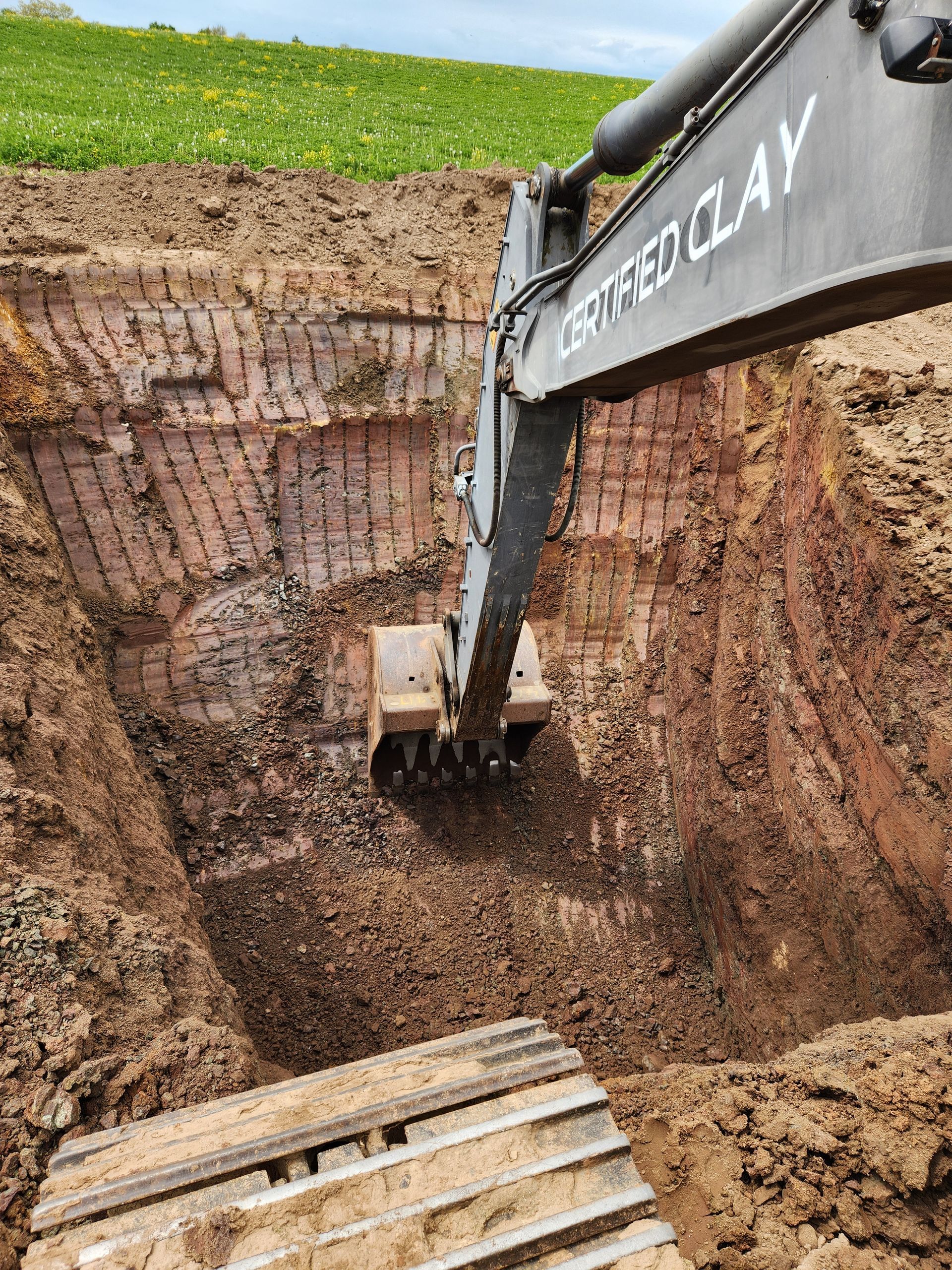An excavator digging a deep trench in brown, muddy soil. The machine's arm and bucket are visible, along with part of its track.