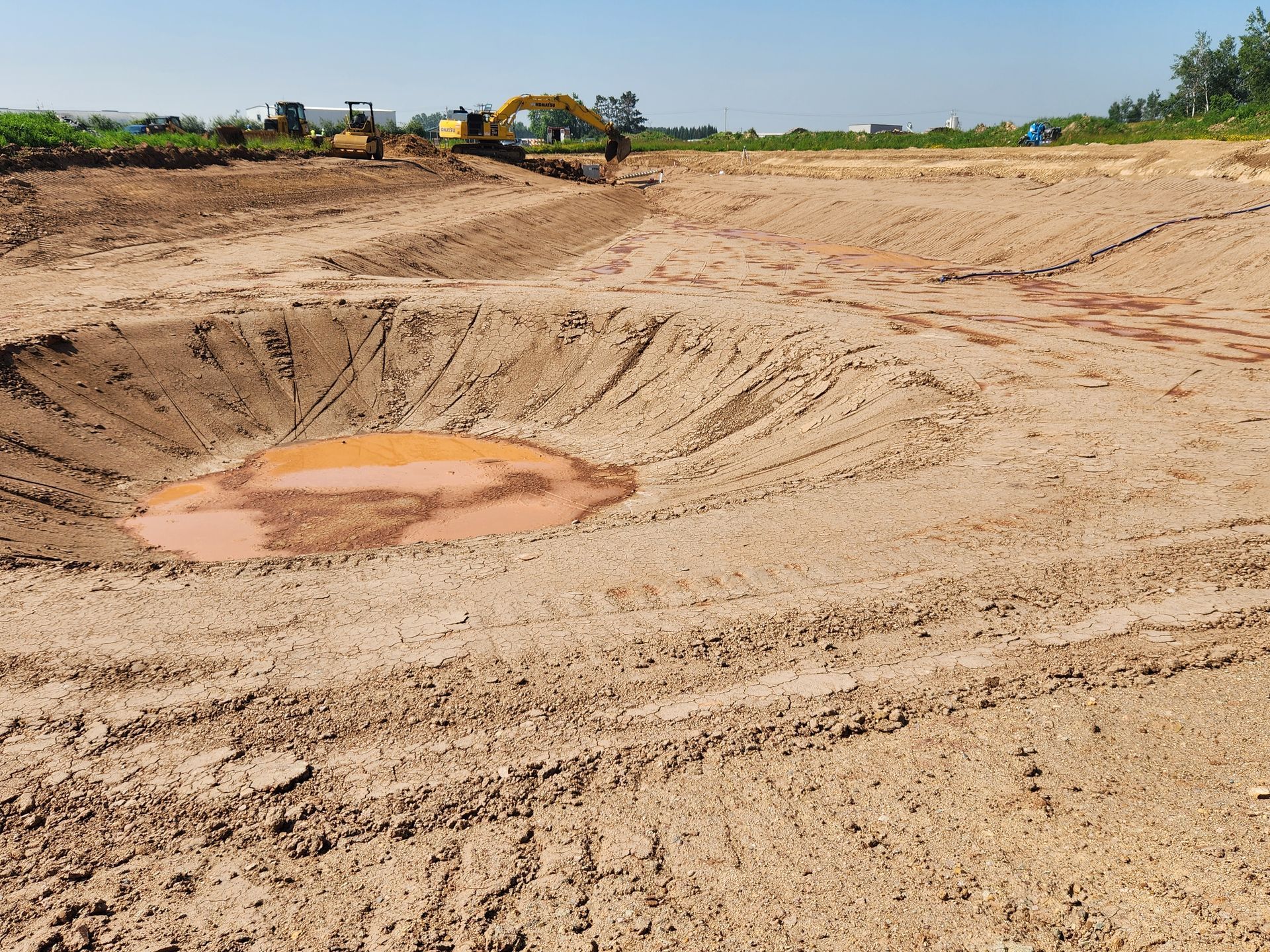 An excavator digs into brown earth, creating a basin that has collected reddish water. Construction site in a sunny setting.