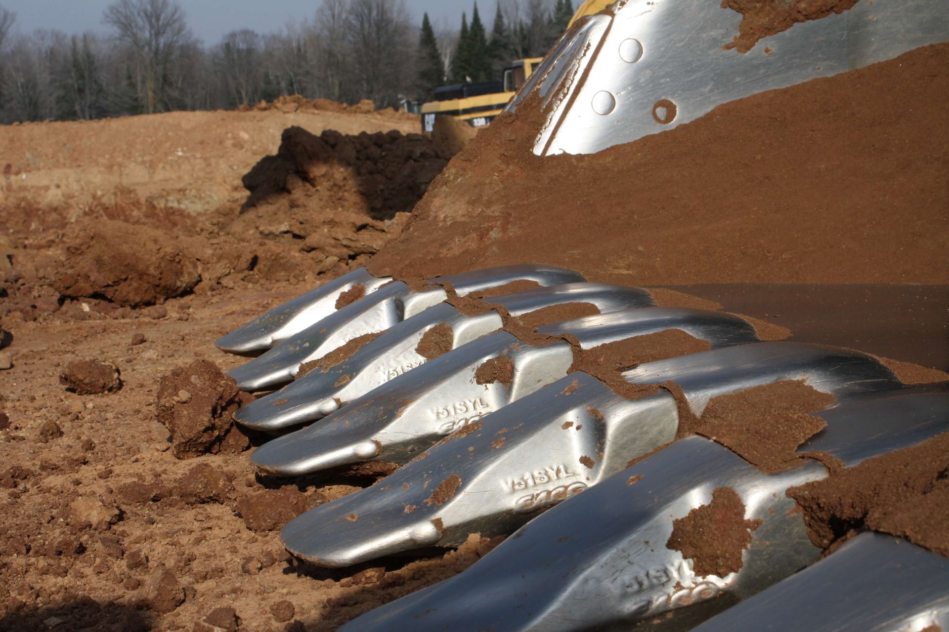 Close-up of excavator bucket teeth digging into brown soil on a construction site.