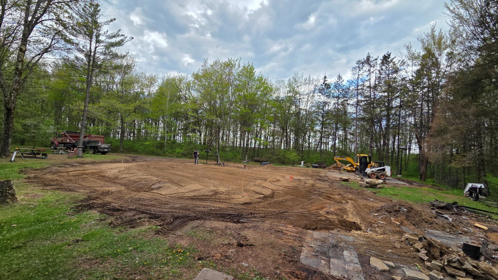 Construction site clearing land surrounded by trees under a cloudy sky. Dirt, machinery, and workers are visible.