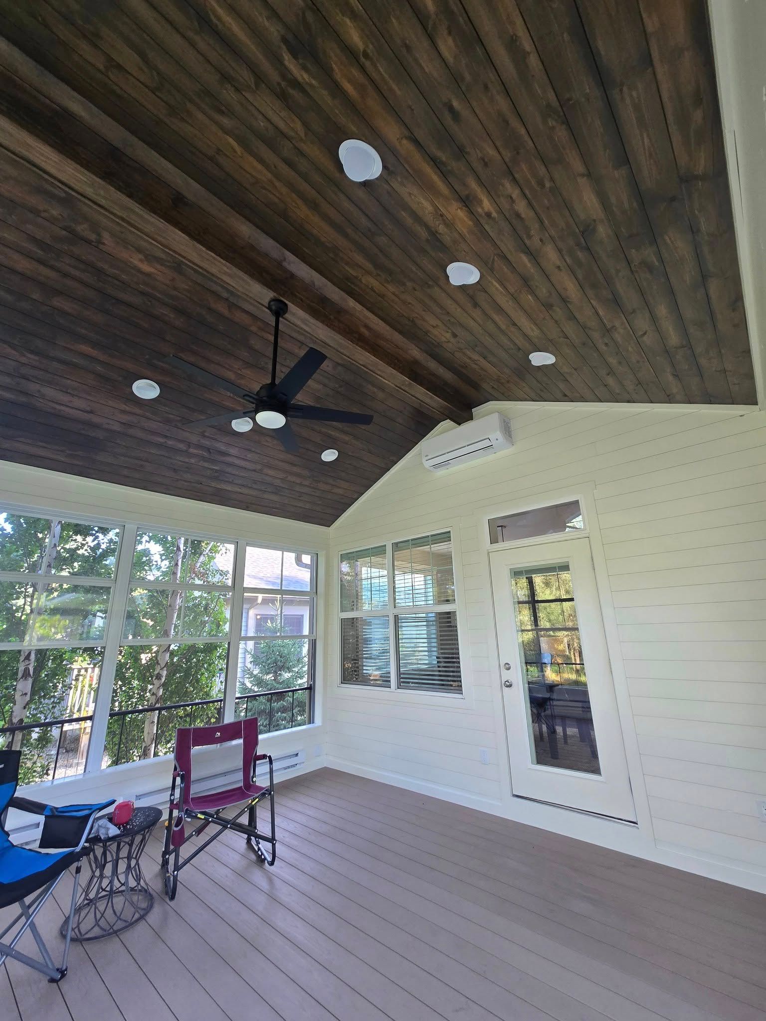 Screened porch with wood ceiling and white walls, furnished with chairs and a ceiling fan.
