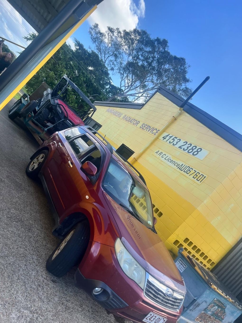 Red Subaru towing a car on a trailer, parked near a yellow building with text— Bundaberg Radiator Service in Bundaberg East, QLD