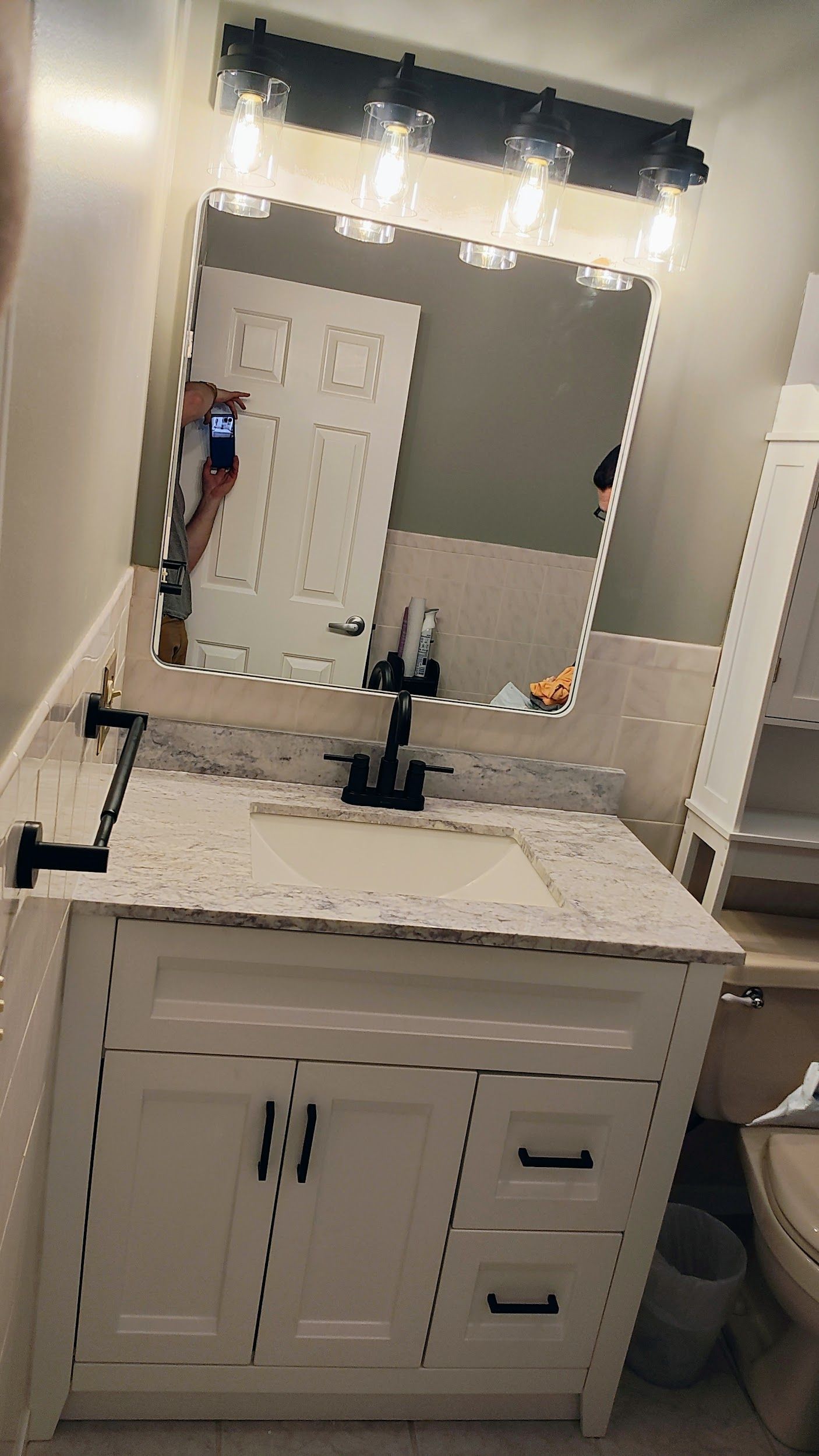 A bathroom vanity with a white cabinet, black hardware, speckled countertop, and a mirror reflecting a person taking a photo.