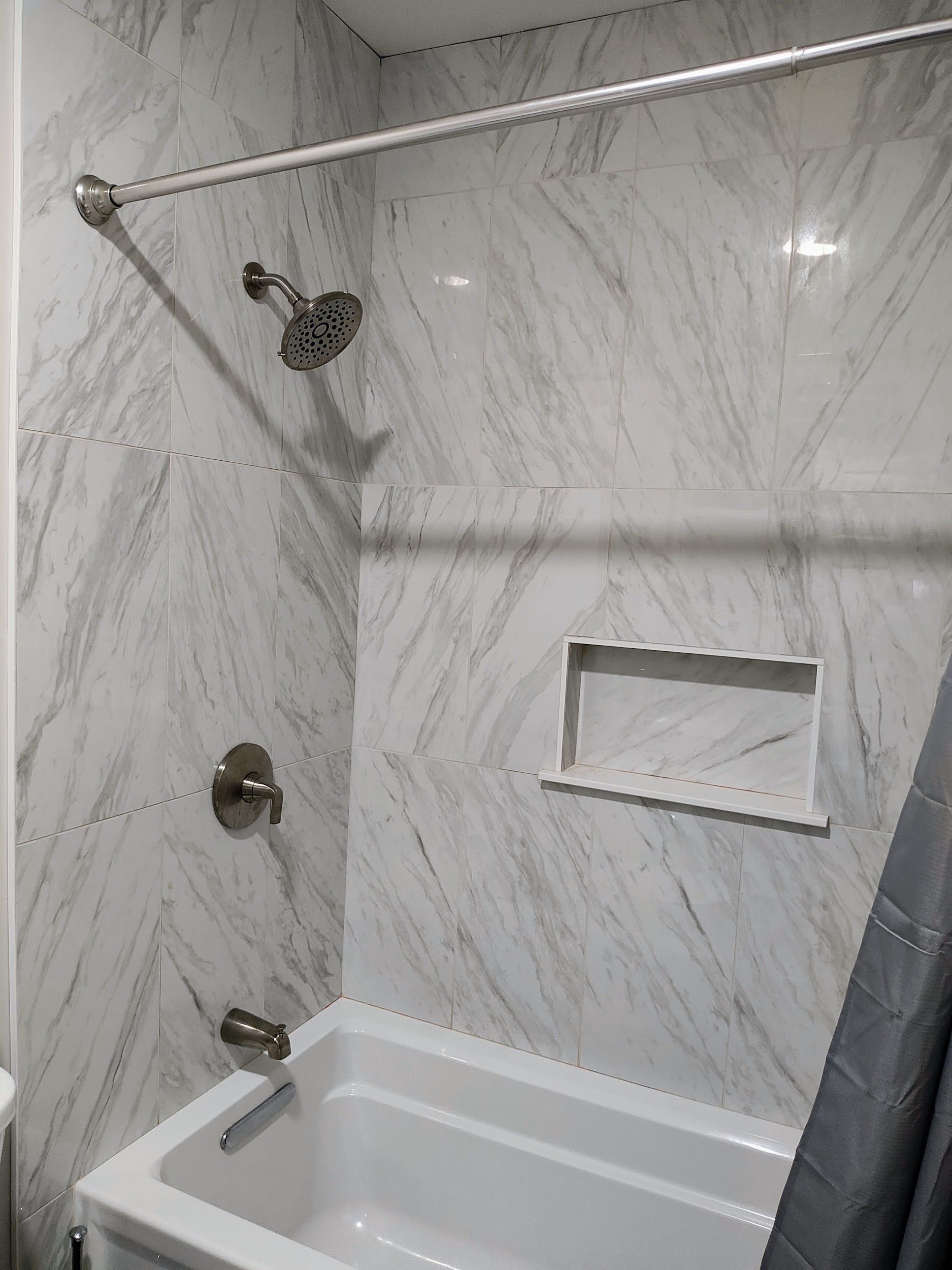 A white marble-tiled shower stall with silver fixtures, a built-in shelf, and a bathtub.