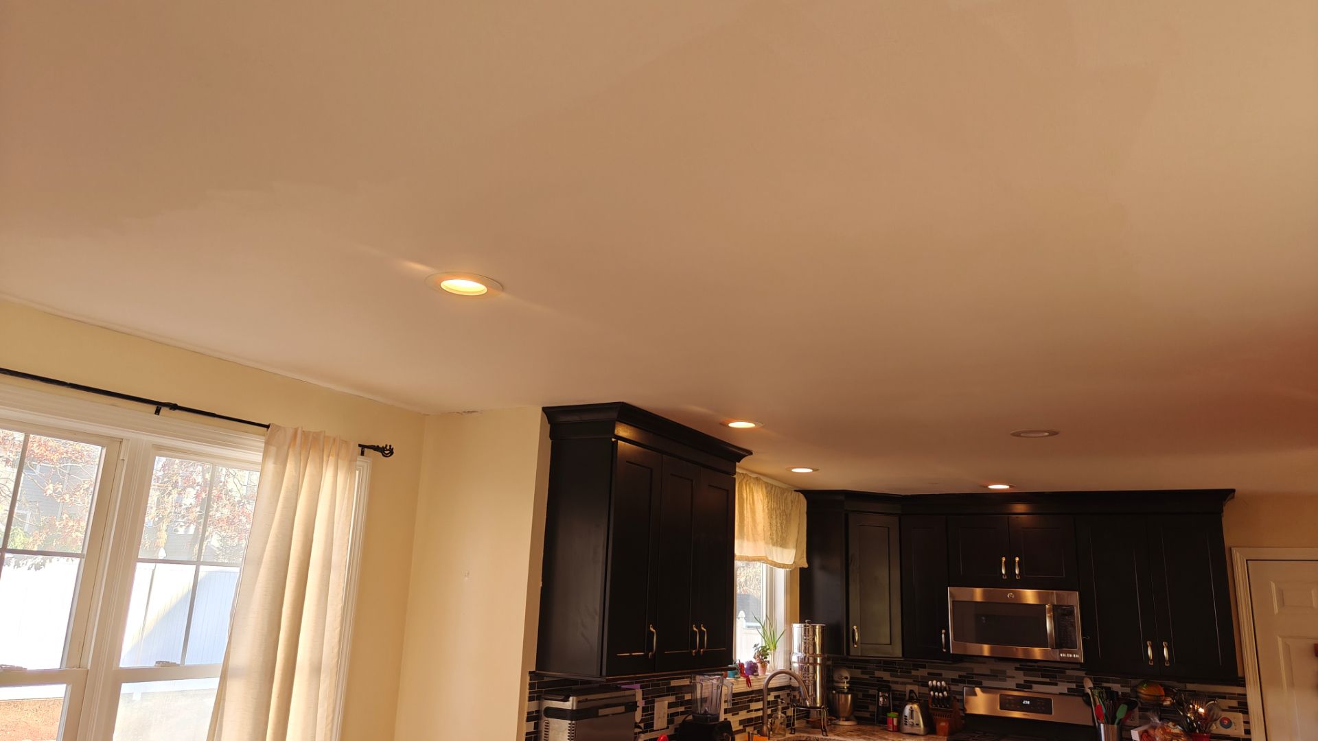 View of a kitchen with dark cabinets, a stainless steel microwave, a window with white curtains, and recessed ceiling lights.