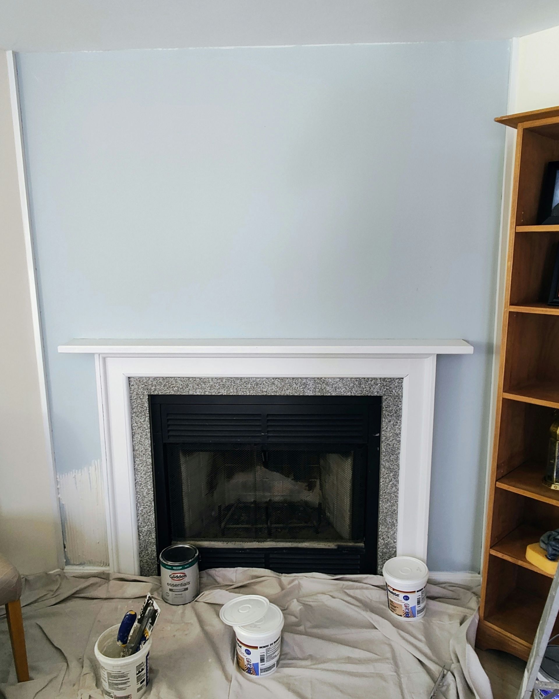 Fireplace with white mantel and blue wall. Paint supplies sit on a drop cloth in front. Wooden bookshelf to the right.