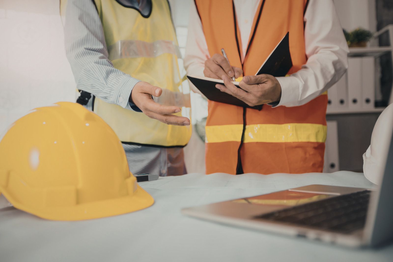 Two people in safety vests reviewing notes near a laptop and yellow hard hat in an office setting.