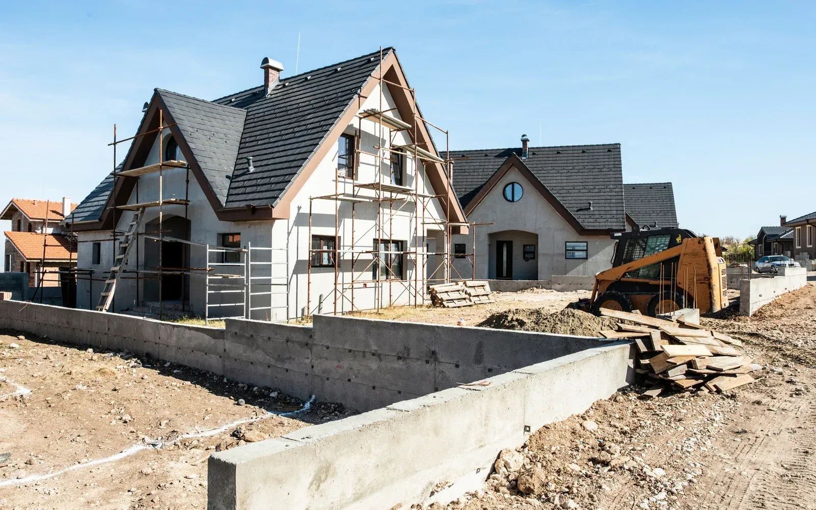 Houses under construction, scaffolding, a backhoe, dirt, concrete barriers, and a bright blue sky.
