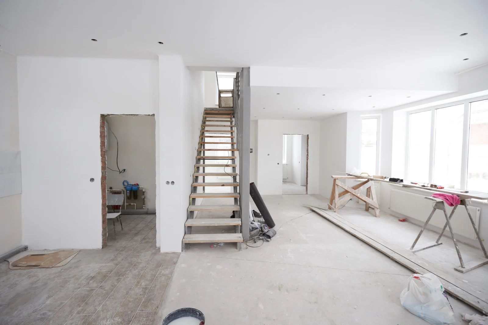 Interior of a house under renovation, with staircase, unfinished floors, and white walls.