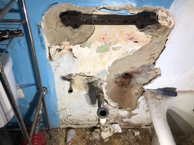 Exposed wall area with pipes, bracket, and peeling paint, with blue and white colors, in a utility setting.