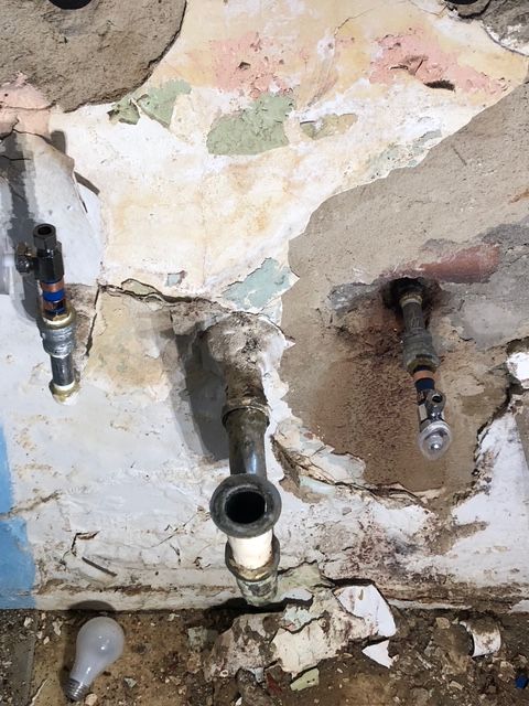 Exposed plumbing pipes in a wall, with peeling paint and a light bulb on the floor.