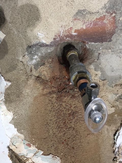 A plumbing fixture with a black shower head extends from a hole in a damaged wall.