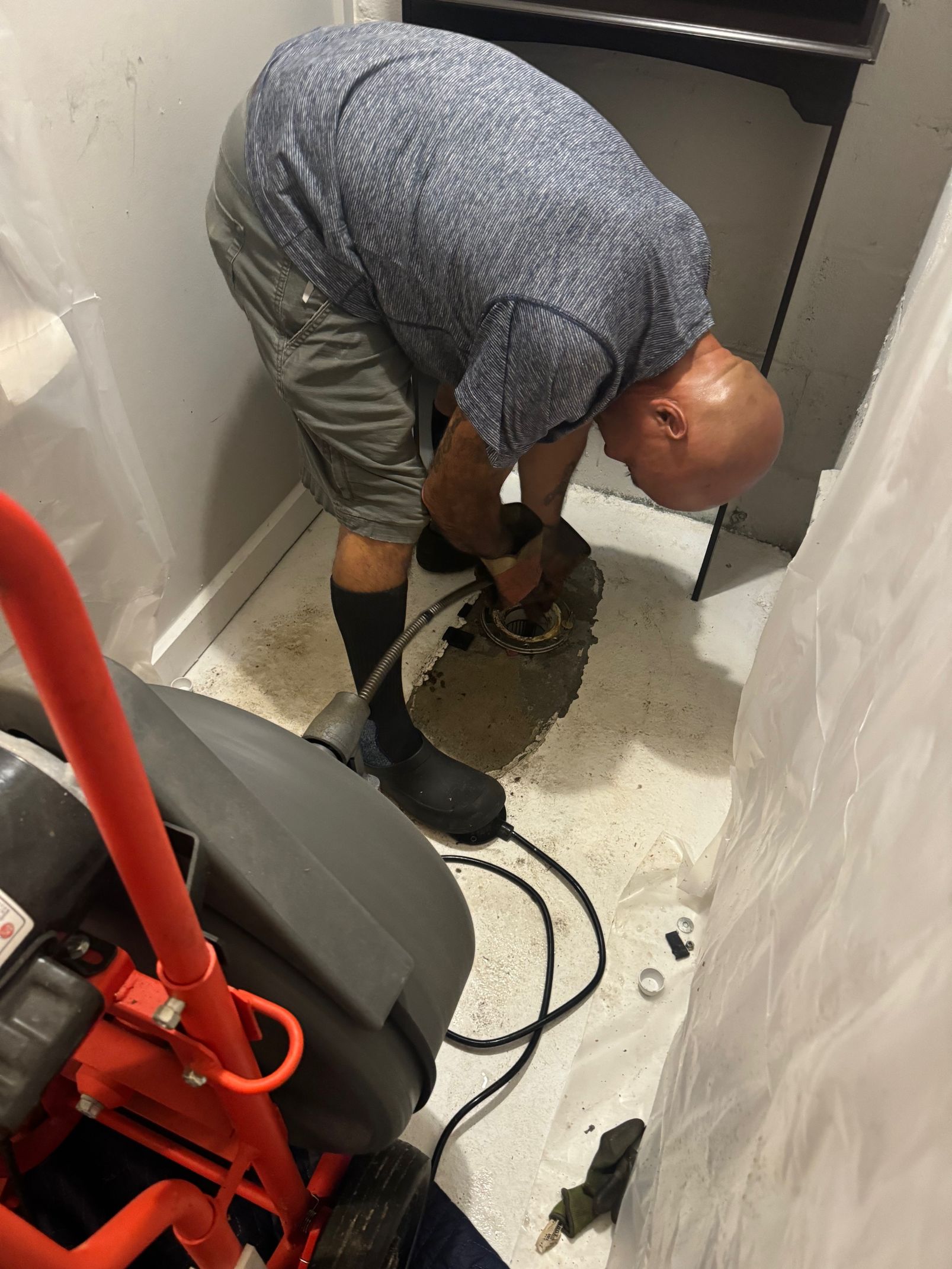 Man in work clothes near a plumbing tool, inspecting a drain in a small room with plastic sheeting.