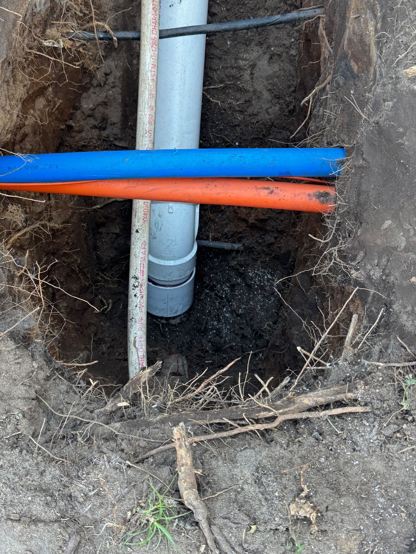 A square hole in the ground reveals grey and colored pipes, roots, and a pole.