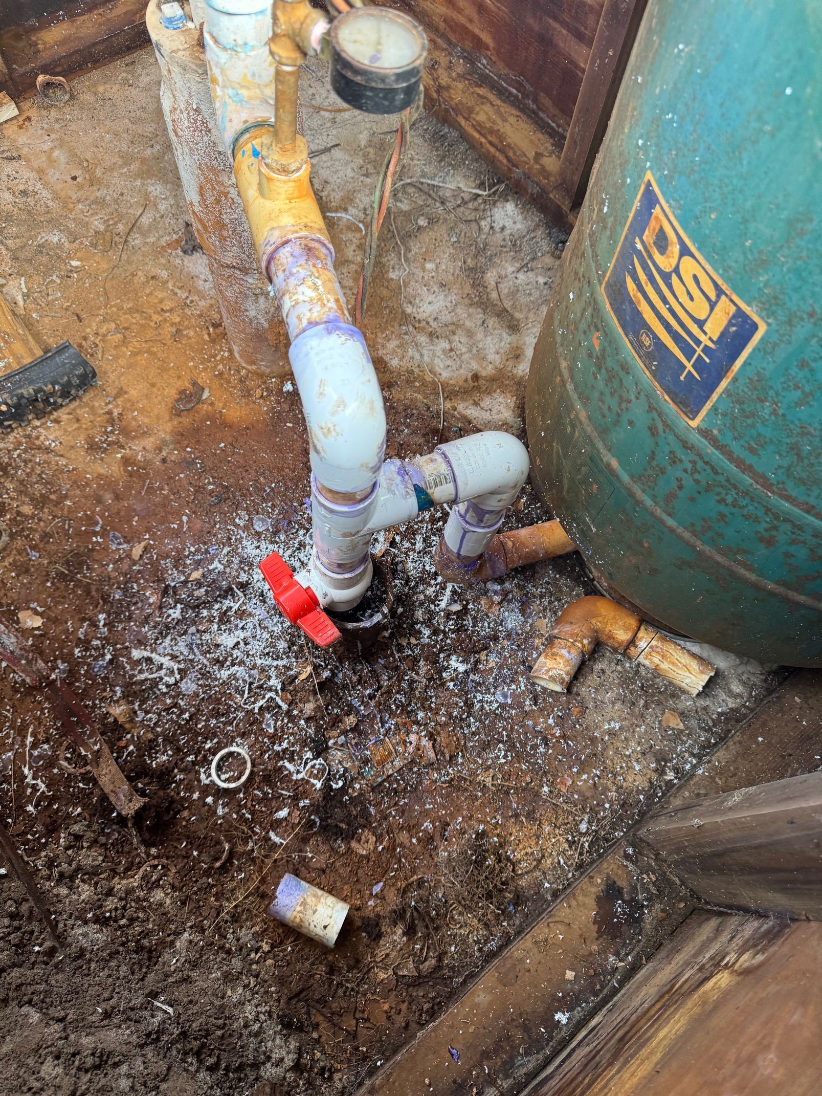Plumbing system near a green water tank. White PVC pipes, red valve, and water on the ground.