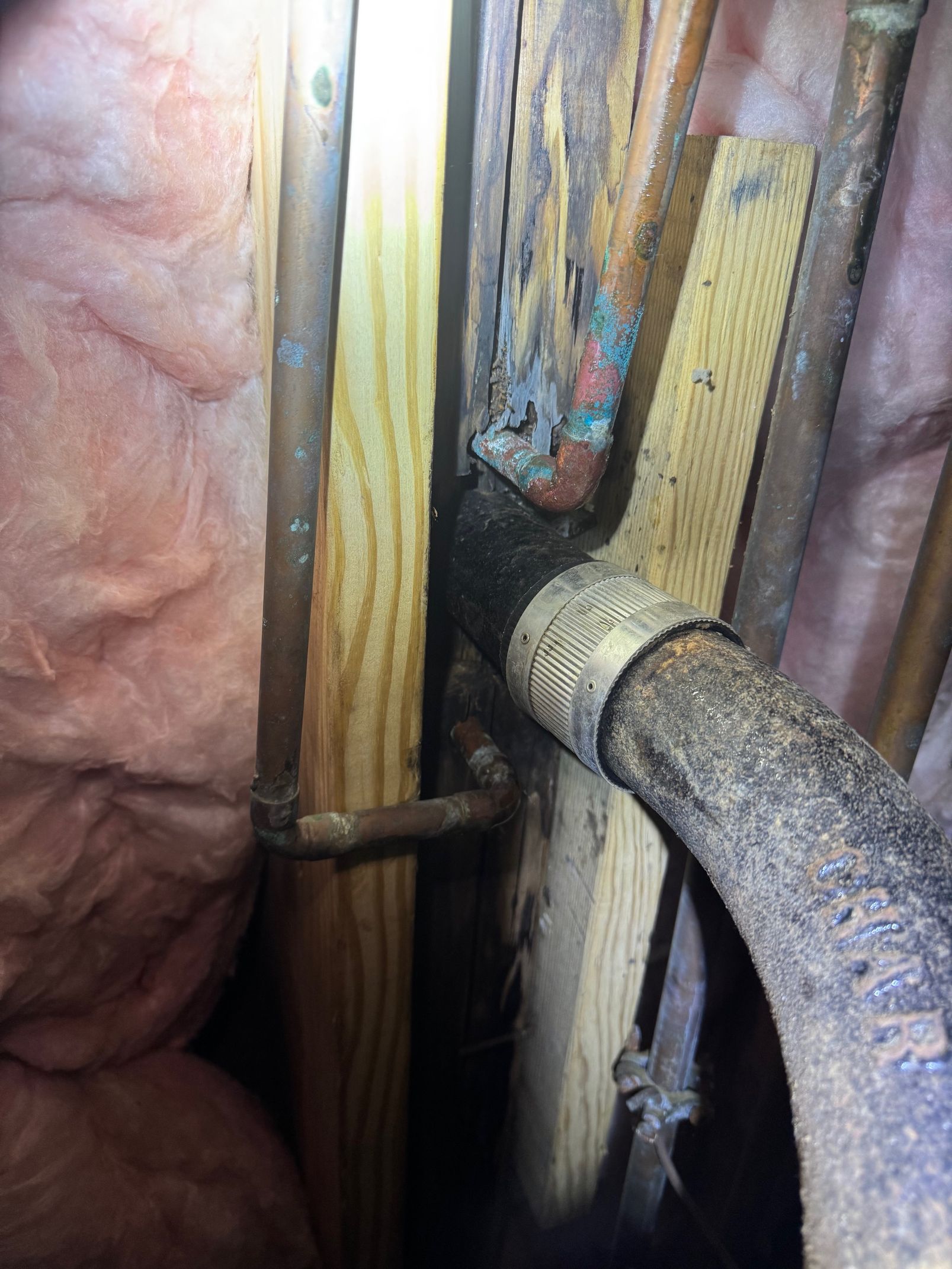 Pipes in a wall cavity, with insulation visible. Copper pipes show corrosion, near a black pipe and wood supports.