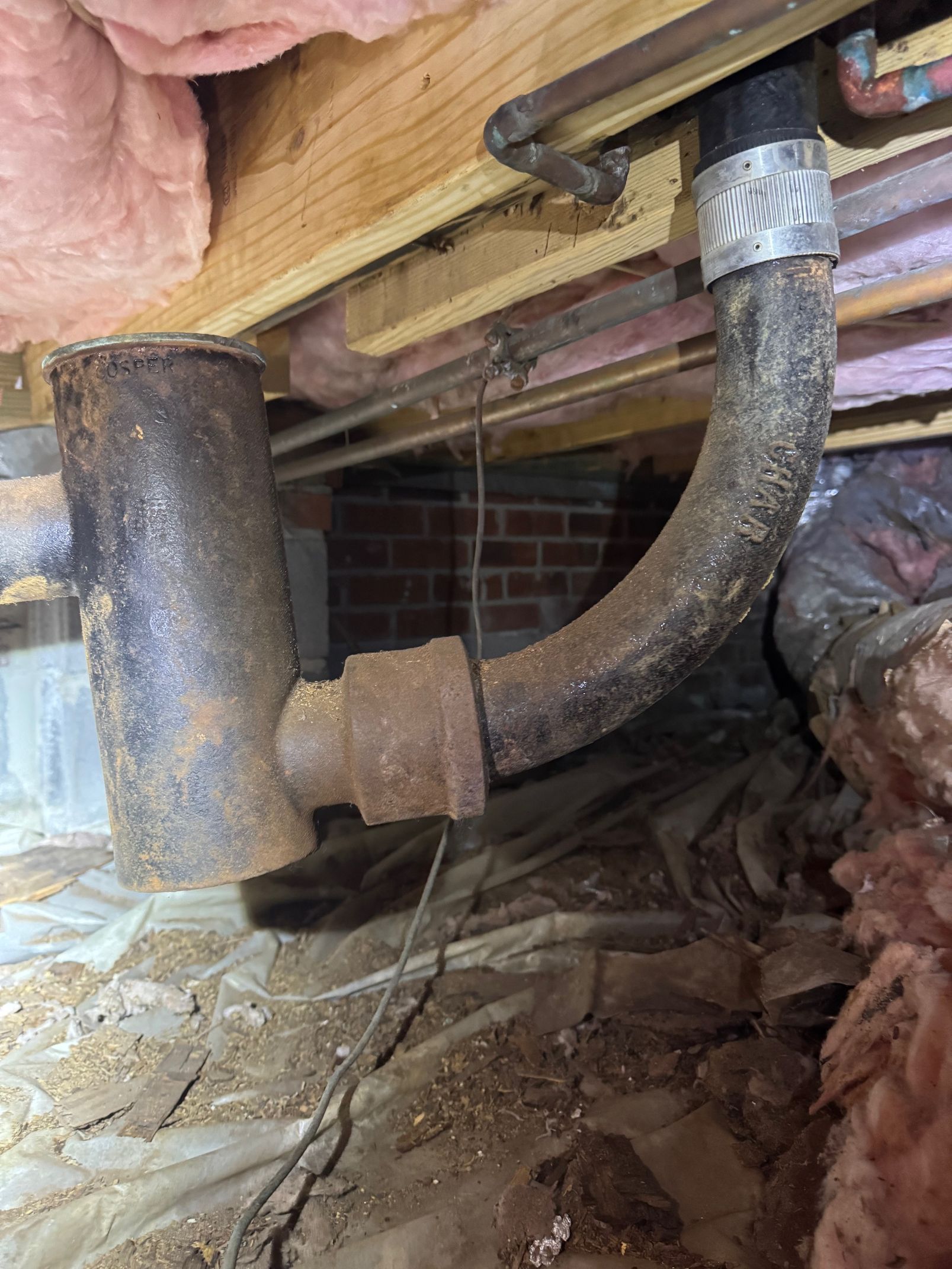 Black cast iron plumbing pipes in a crawl space, connected with a curved drainpipe near insulation and wooden beams.
