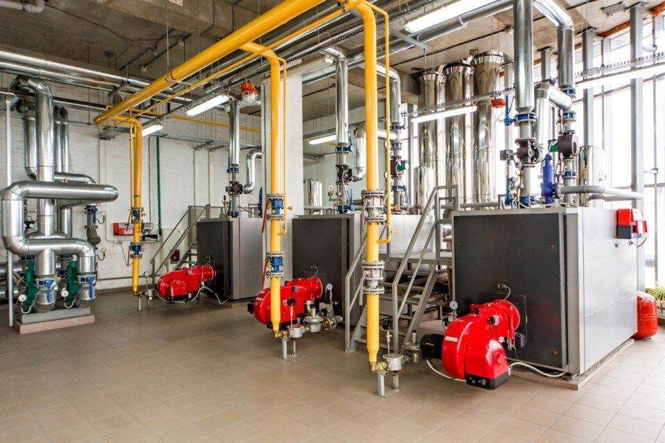 Industrial boiler room with yellow and silver pipes, gray machinery, red burners, and windows.