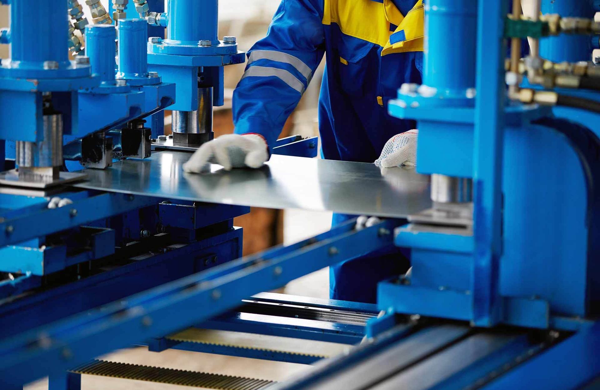 A worker in a blue and yellow uniform uses gloved hands to guide a flat sheet of metal through a blue industrial machine.