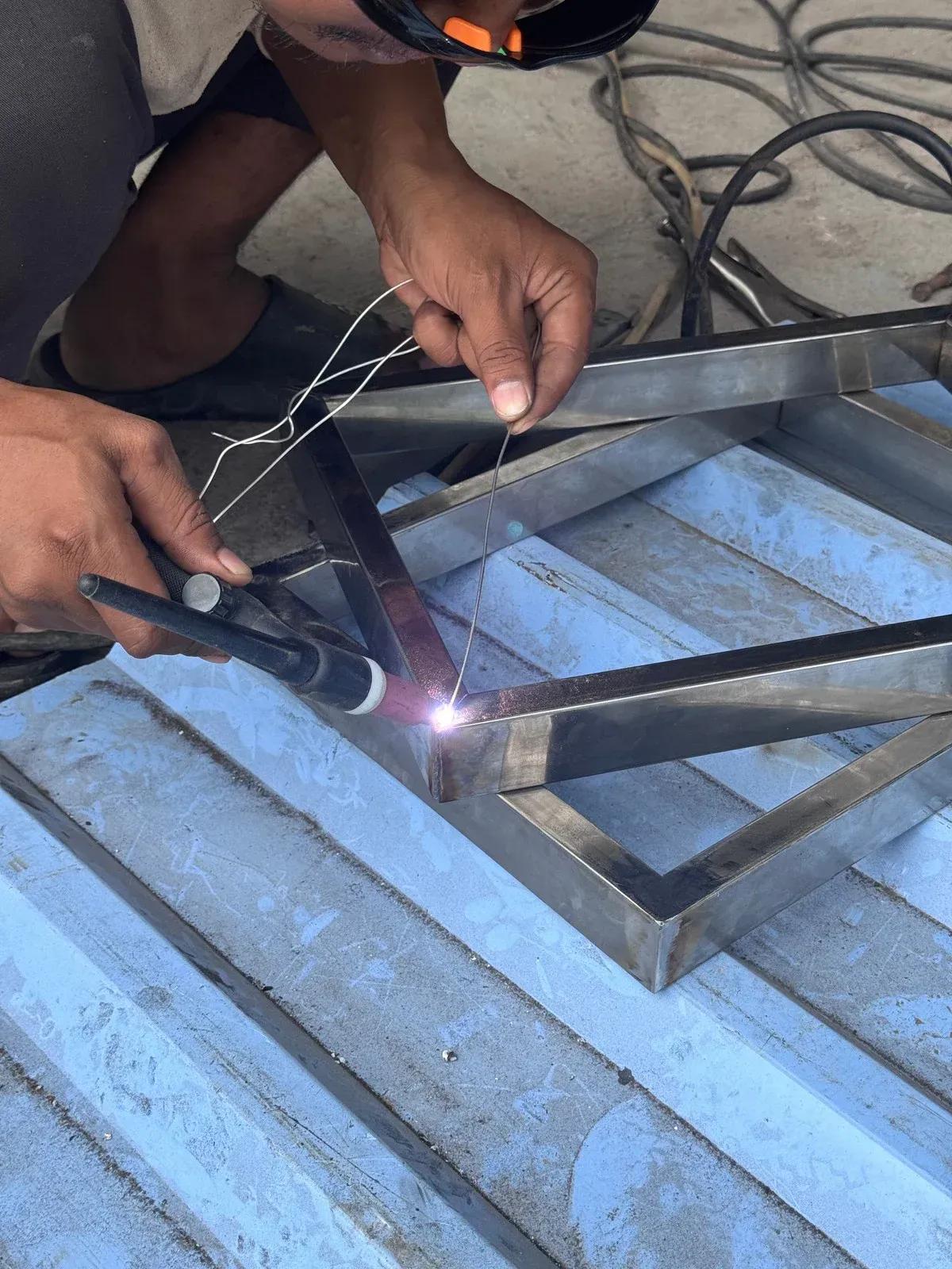 A person welding metal frame outdoors, wearing a welding mask, with sparks visible.