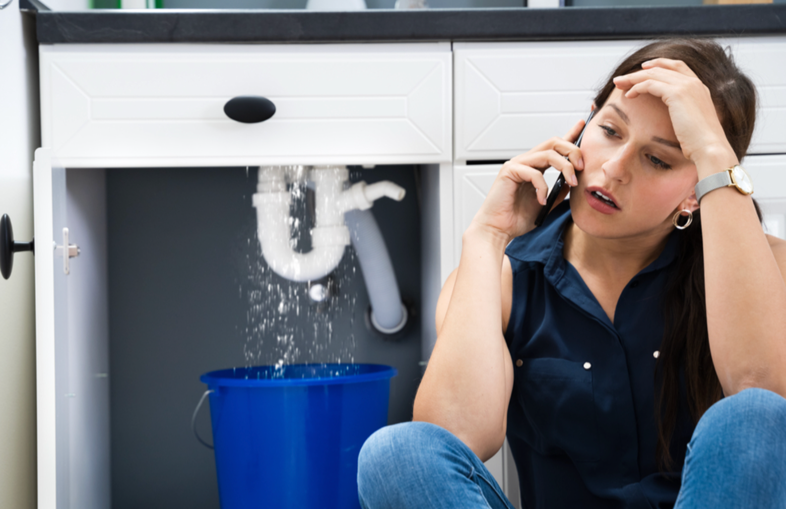 Woman on phone, distressed, with leaking sink pipe, blue bucket collecting water under cabinet.