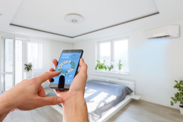 Hands holding a smartphone showing a smart home app in a bright bedroom