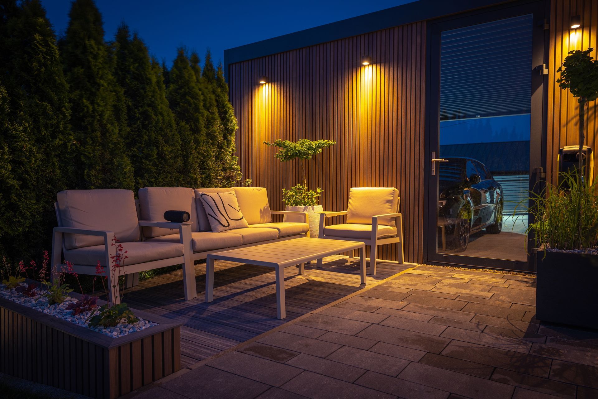 Modern patio lounge with white sofas, warm lighting, and potted plants at dusk