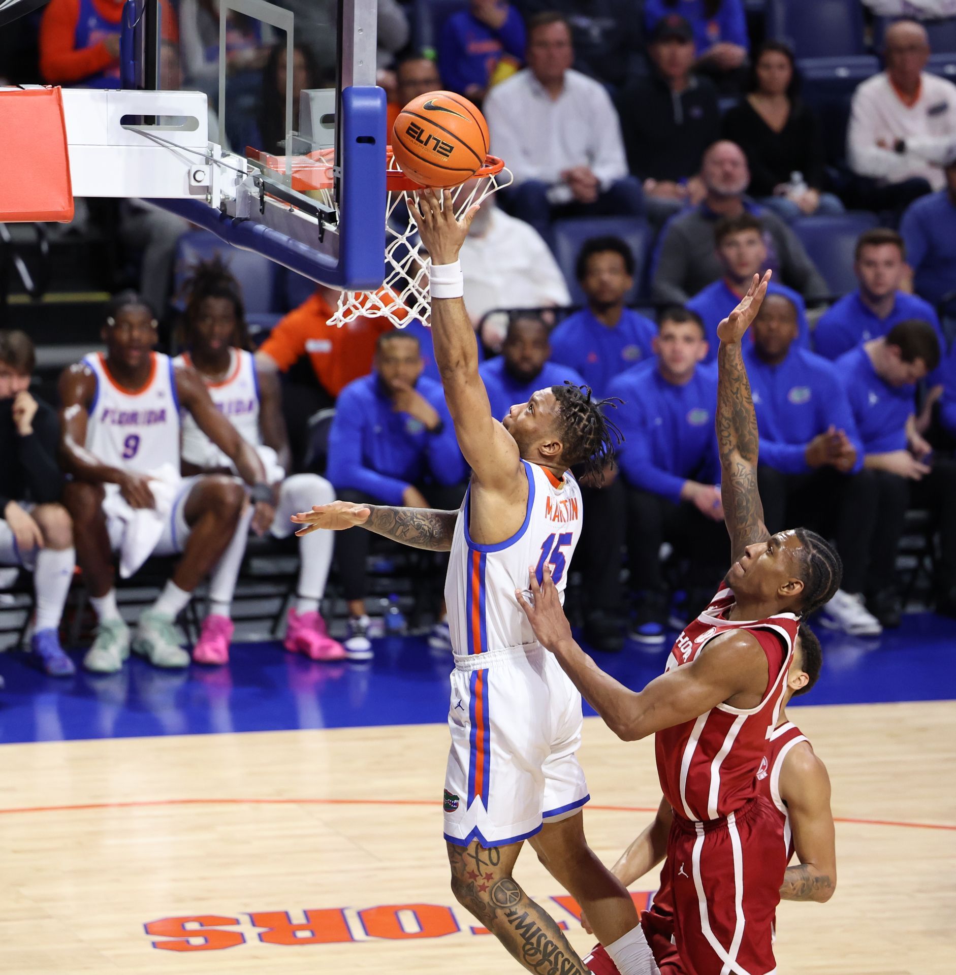 Basketball player in blue jersey shoots a layup, another player defends, crowd in background.
