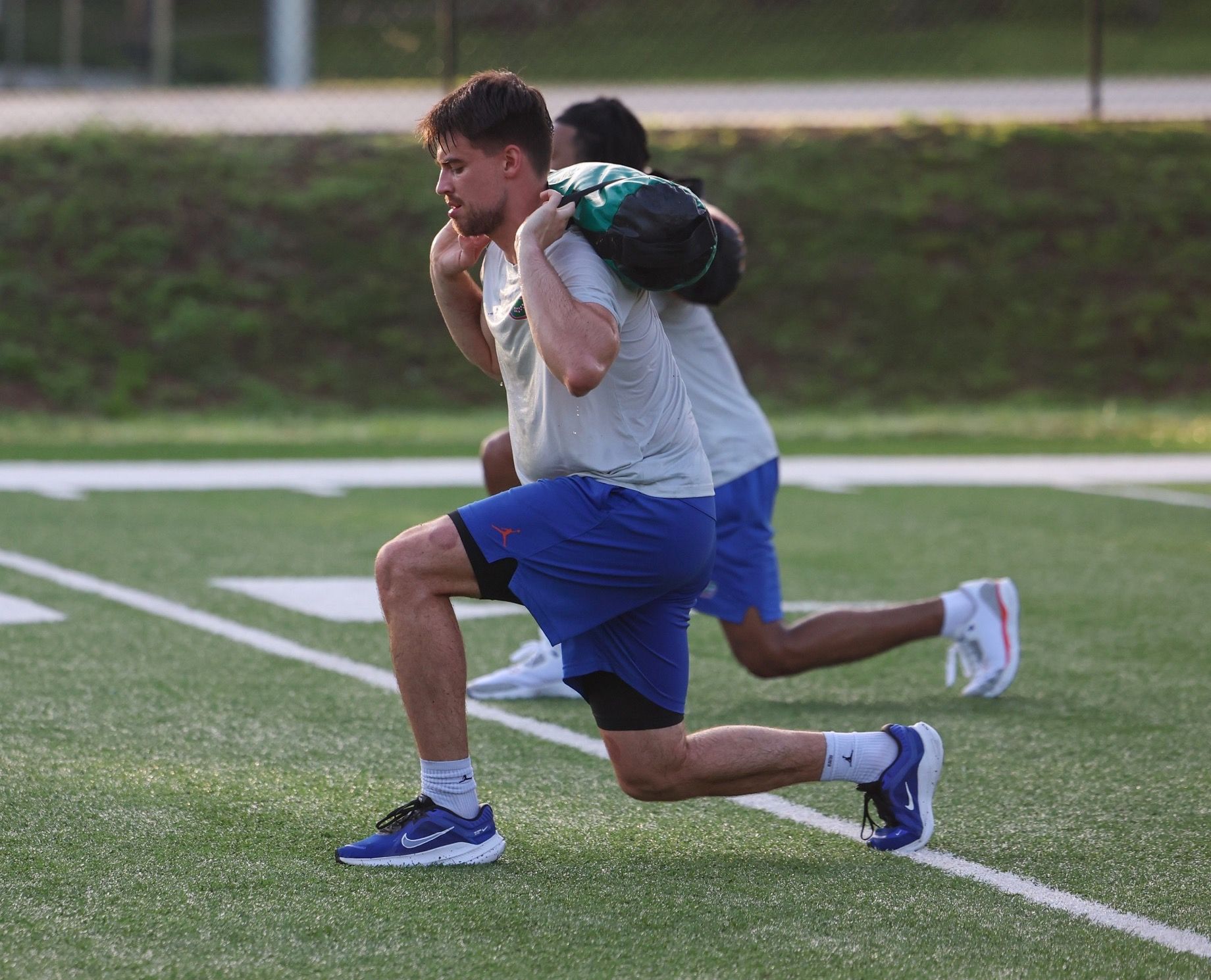 Two men are doing lunges on a field with a bag on their backs.