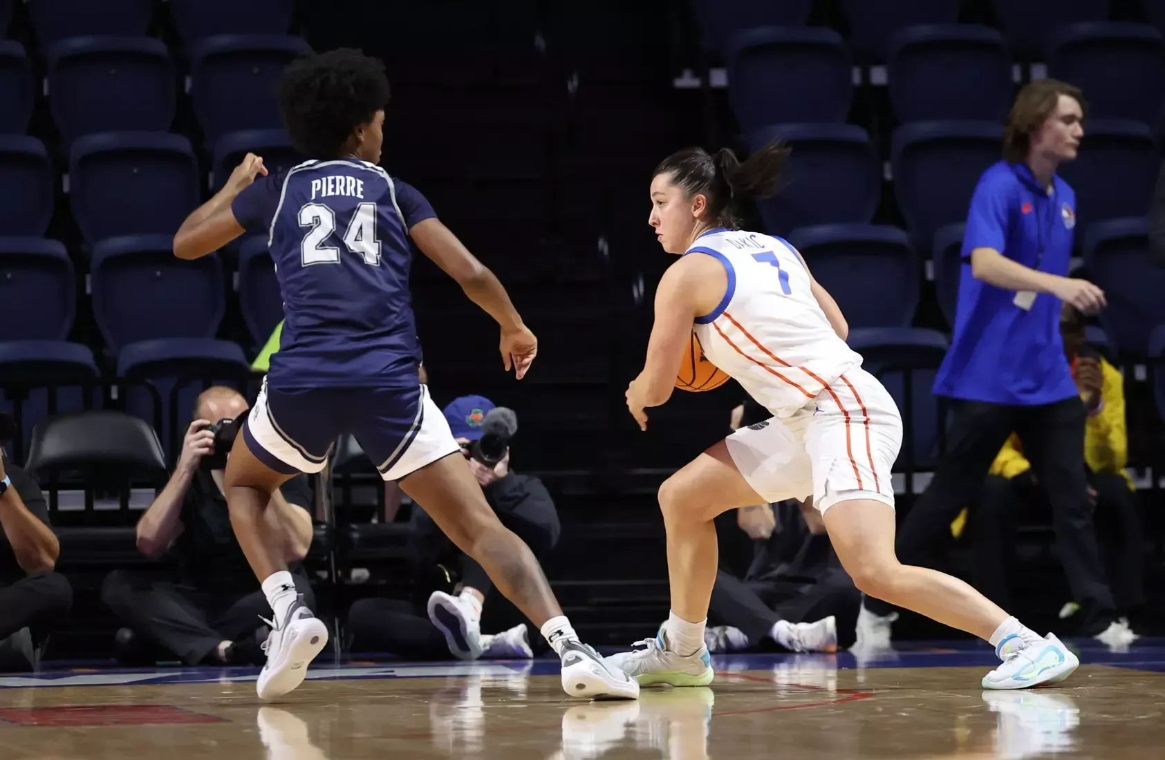 Basketball player dribbles ball, guarded by opponent on court. Spectators in background.