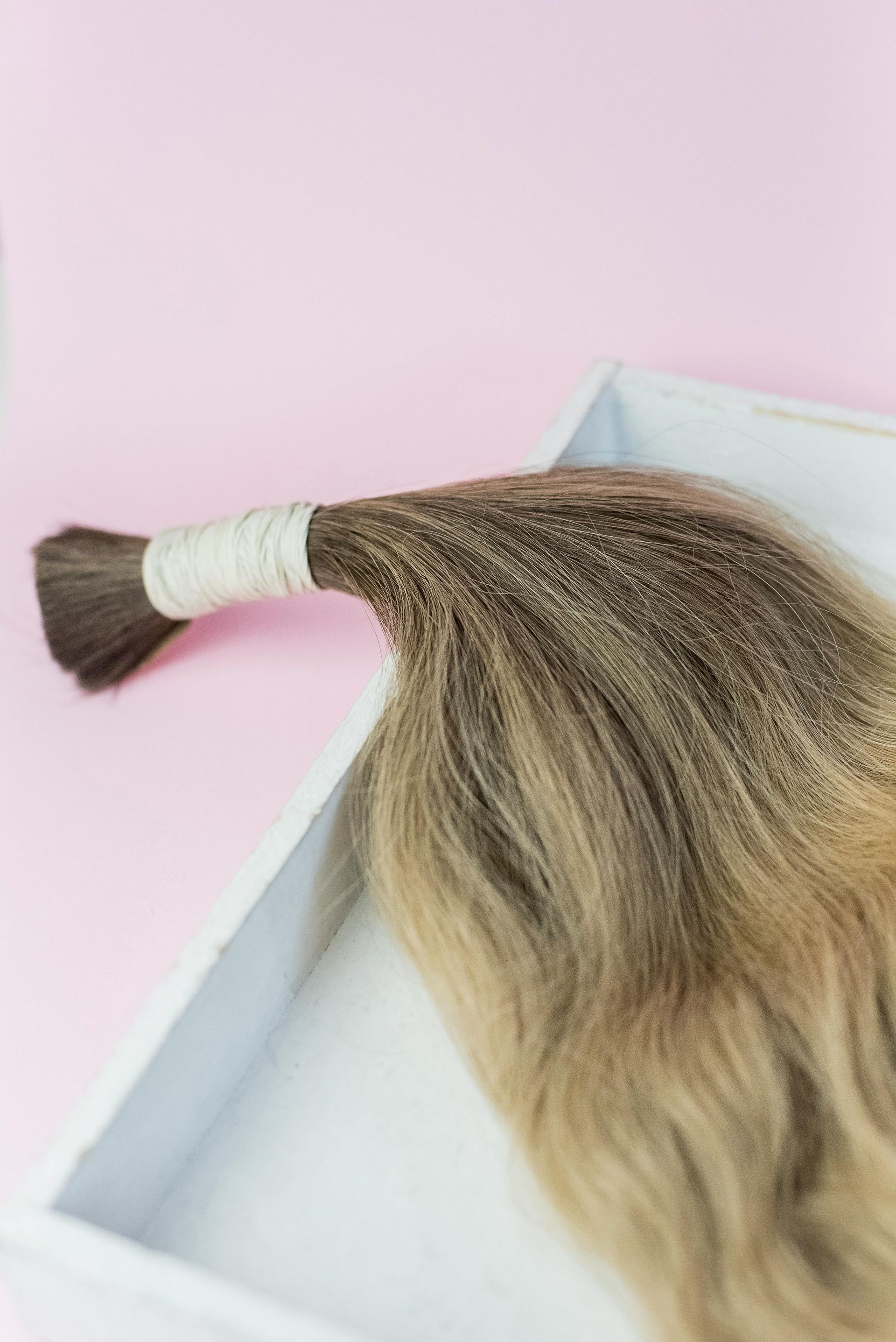 A bundle of brown and blonde hair tied with white string, resting in a white tray against a pink backdrop.
