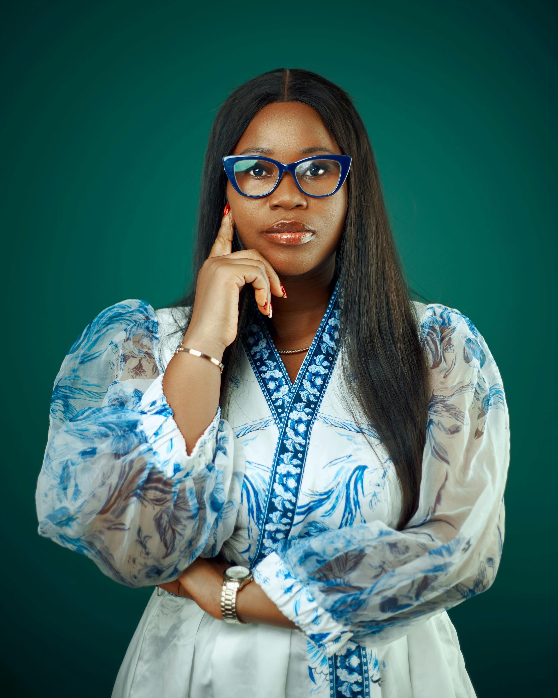 Woman with glasses, hand on chin, wearing a white and blue floral robe, posing against a green backdrop.